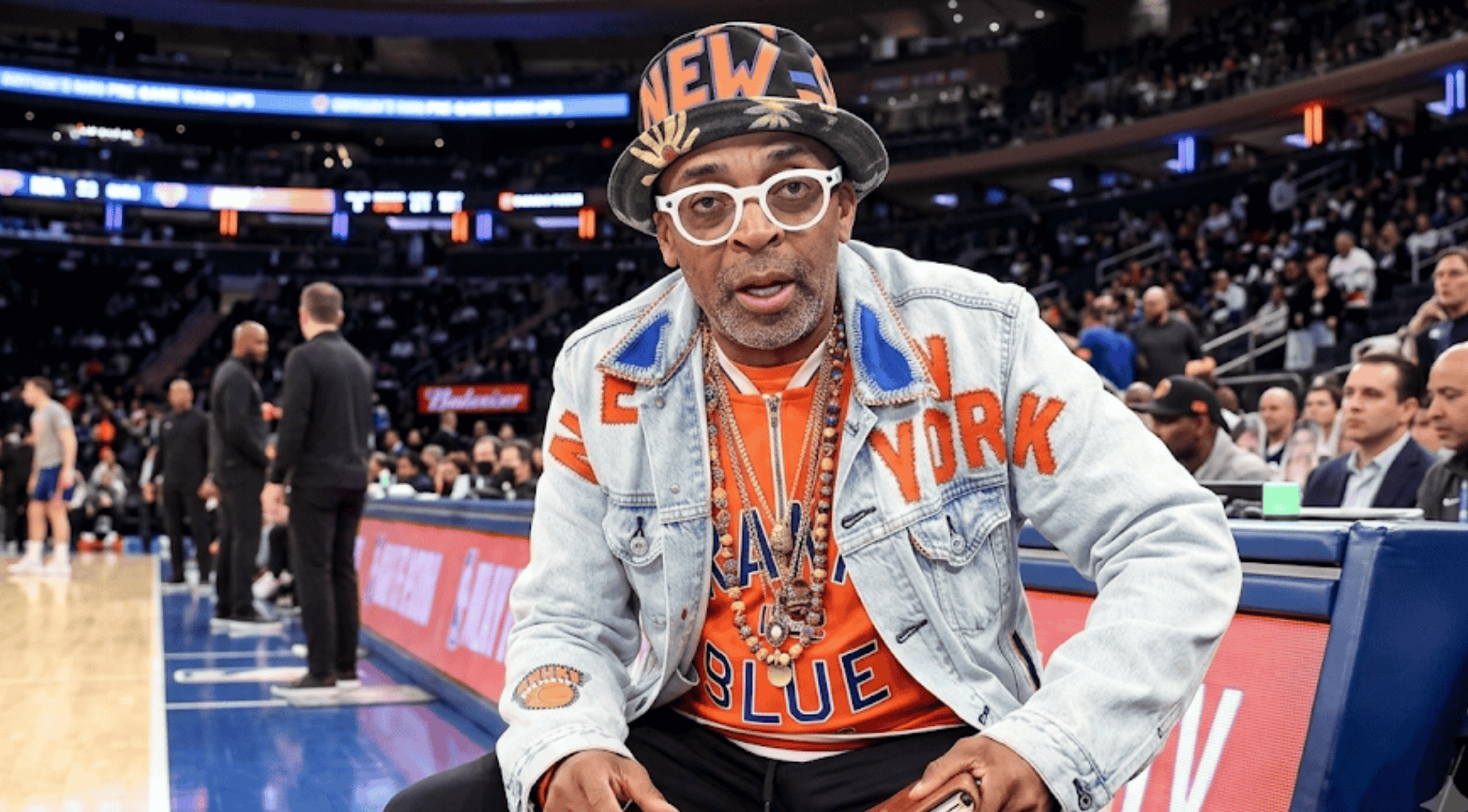 Spike Lee sitting courtside wearing his custom light-wash denim trucker jacket featuring orange "NEW YORK" patchwork appliqué and a circular Knicks logo on the sleeve.